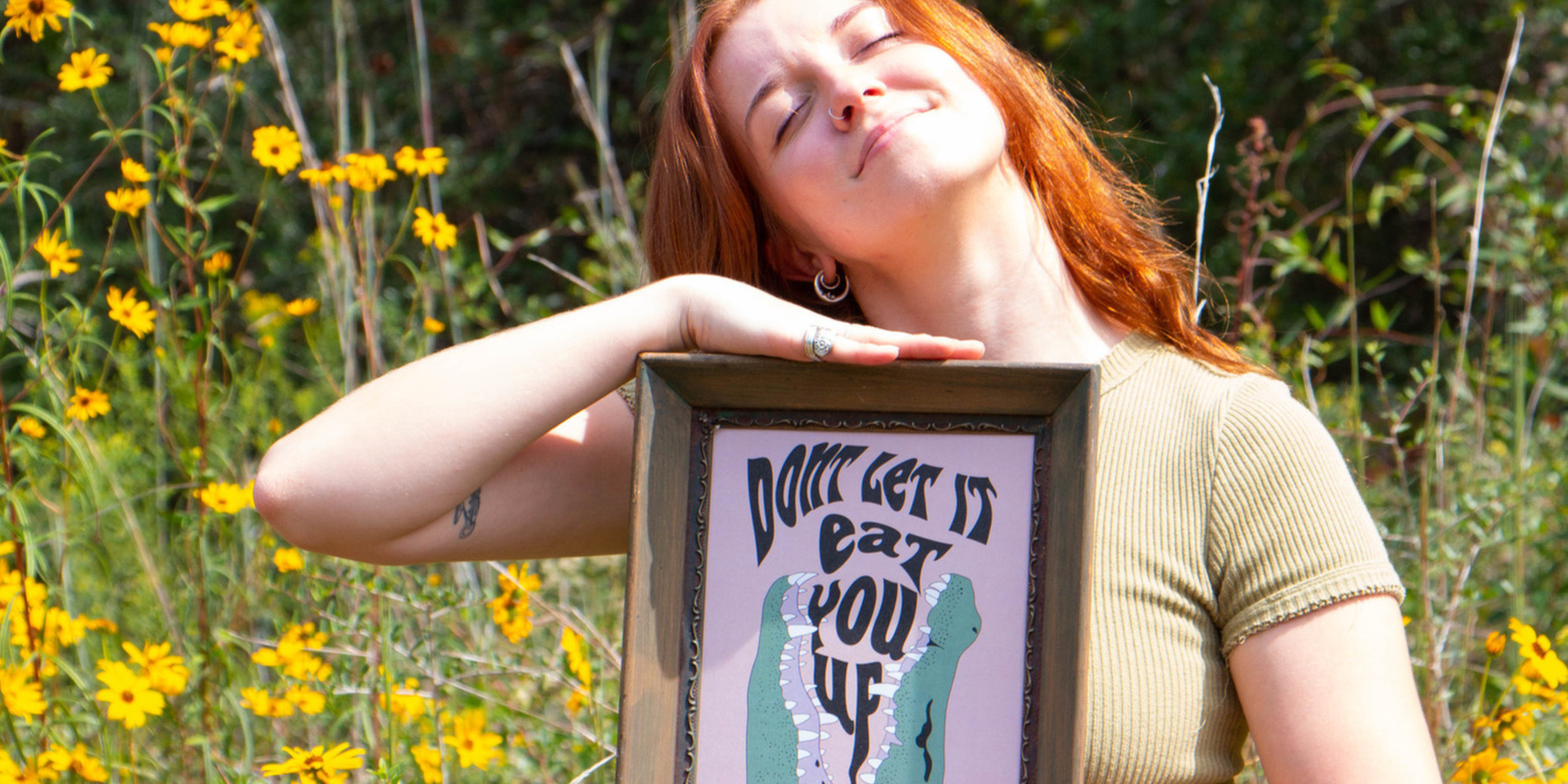 Photo of Louisiana artist, Tristen Rolling, holding a framed print of her digital illustration titled, "don't let it eat you up," which features a Louisiana alligator eating the phrase that the illustration gets its namesake from. She is standing in a field of Louisiana wildflowers looking up at the sunlight. 