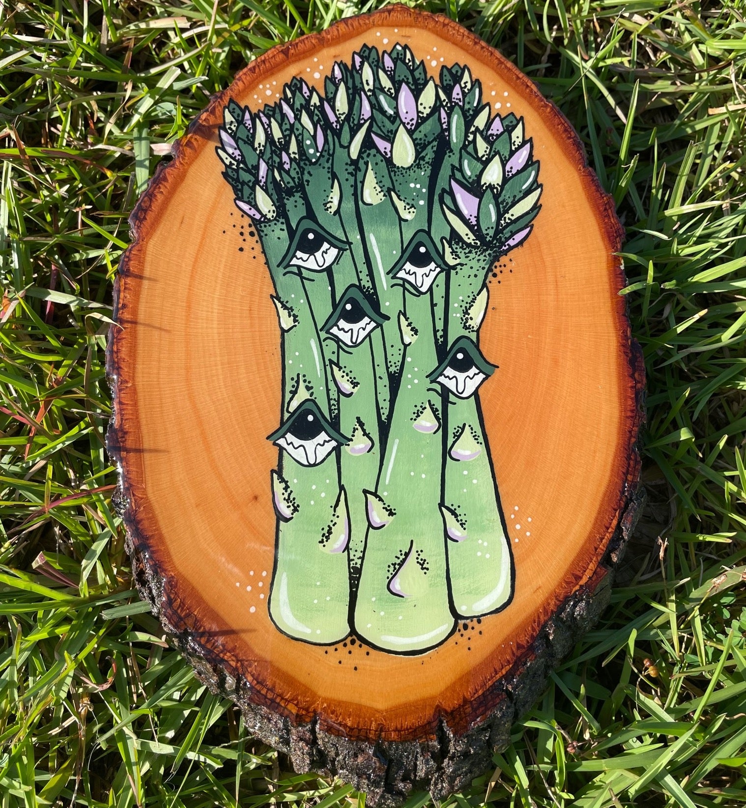 A photo of Louisiana artist, Tristen Rolling's original acrylic painting of a bunch of asparagus on a round slice of oak wood. The painting is coated in a layer of epoxy resin and reflects Tristen's love for Louisiana farmers markets.  